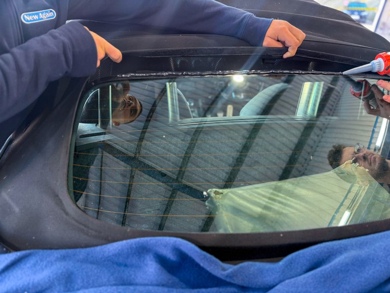 Two New Again technicians applying sealant along the top edge of a convertible glass rear window during a rebond, hood held back and the bodywork covered