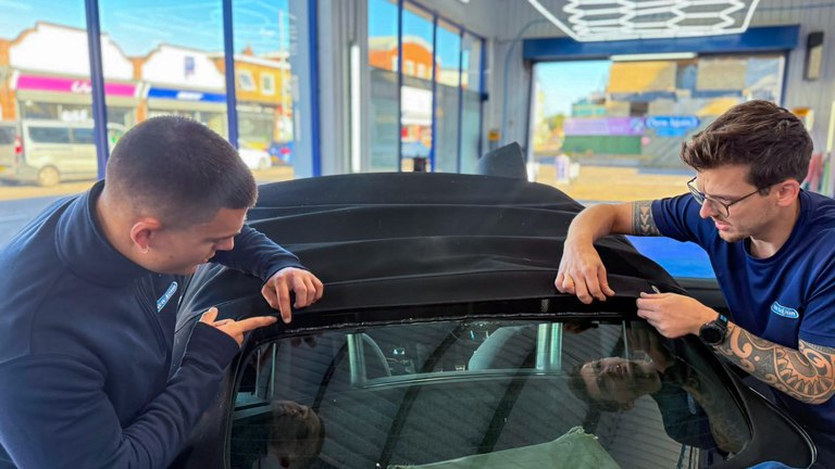 Matt and Alfie cleaning up the bond line where a glass rear window has come away from a black convertible hood, ready for rebonding