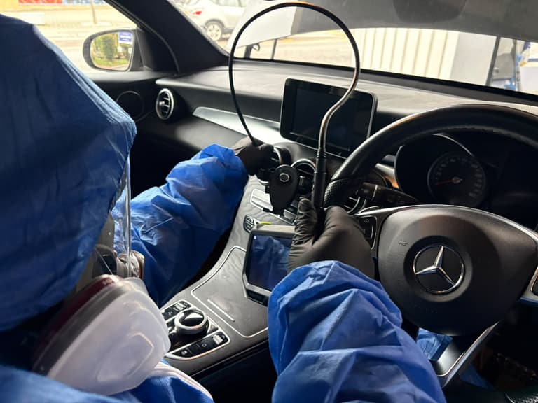 A technician in a coverall and full-face respirator feeding an endoscope into the dashboard air vents of a Mercedes.