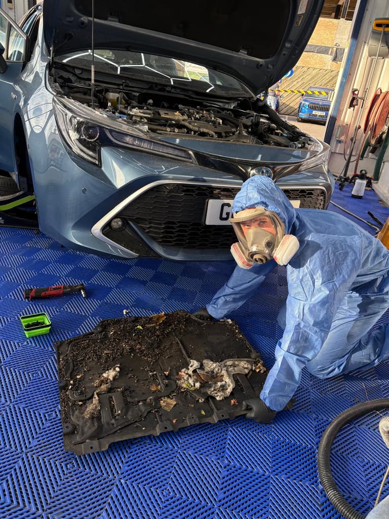 Harry from the New Again team in a hooded coverall and full-face respirator beside a rodent-contaminated engine undertray.